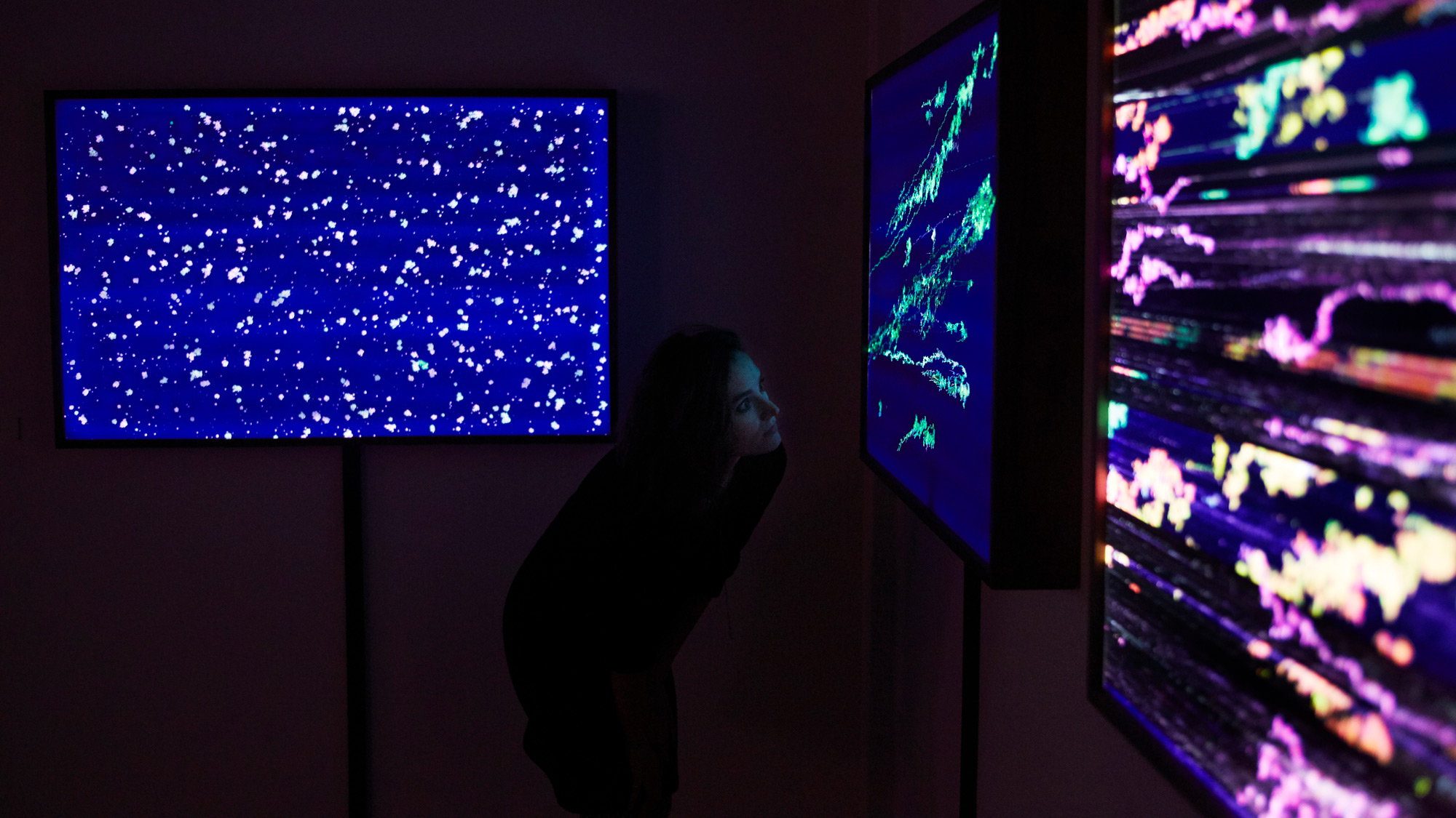 Photograph of a woman in a dark room looking at an illuminated green artwork on a wall, flanked by two other illuminated artworks on either side